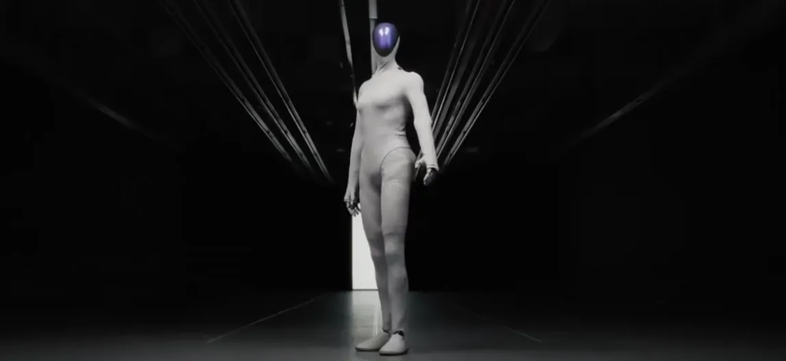 La imagen muestra una figura humanoide de pie en un entorno oscuro, iluminada por una luz que proviene de un fondo brillante. La figura, vestida con un traje ajustado y sin rasgos faciales, tiene una cabeza esférica de color púrpura. Su postura es erguida y su brazo está extendido hacia adelante, sugiriendo una invitación o conexión. La atmósfera es futurista y enigmática, evocando sensaciones de curiosidad y misterio.